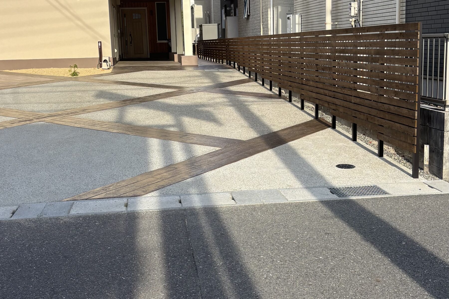 シンプルなベージュの外壁、木材のアクセント、近隣にはレンガ調の淡色建物